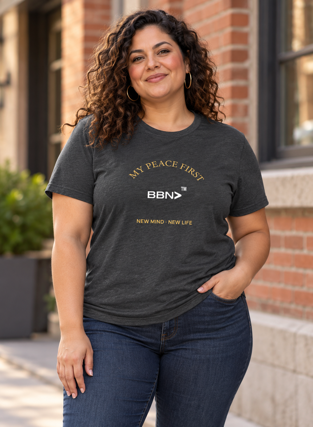 AI styling concept: Woman outdoors wearing a dark gray heather T-shirt with gold “My Peace First” lettering and a white BBN logo.