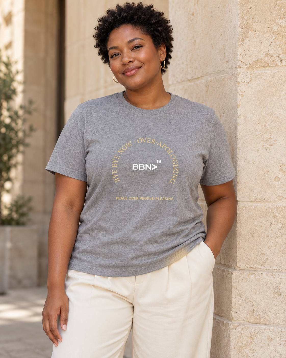 AI styling concept: Woman outdoors wearing an athletic heather gray T-shirt with gold “Over-Apologizing” lettering around a white BBN logo.
