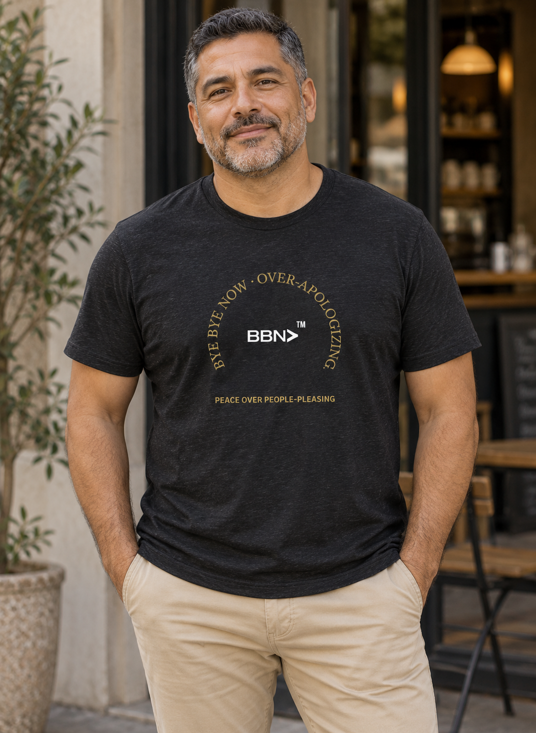 AI styling concept: Man outdoors wearing a black heather T-shirt with gold “Over-Apologizing” lettering around a white BBN logo.