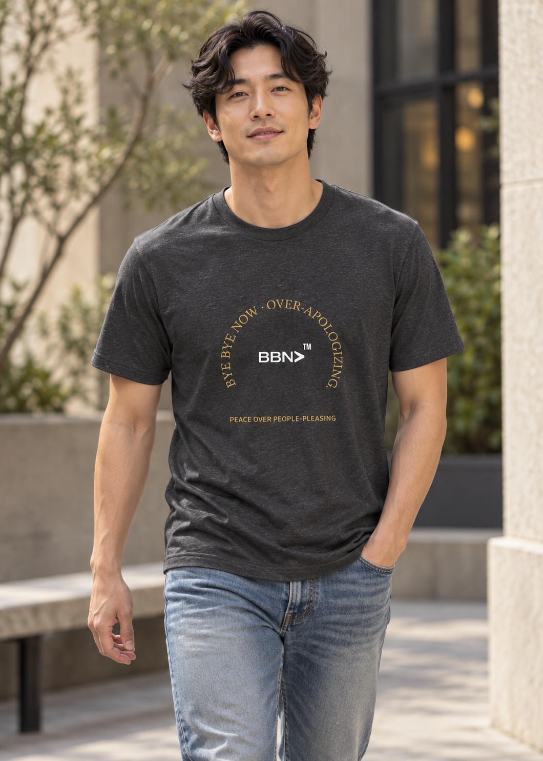 AI styling concept: Man outdoors wearing a dark gray heather T-shirt with gold “Over-Apologizing” lettering around a white BBN logo.
