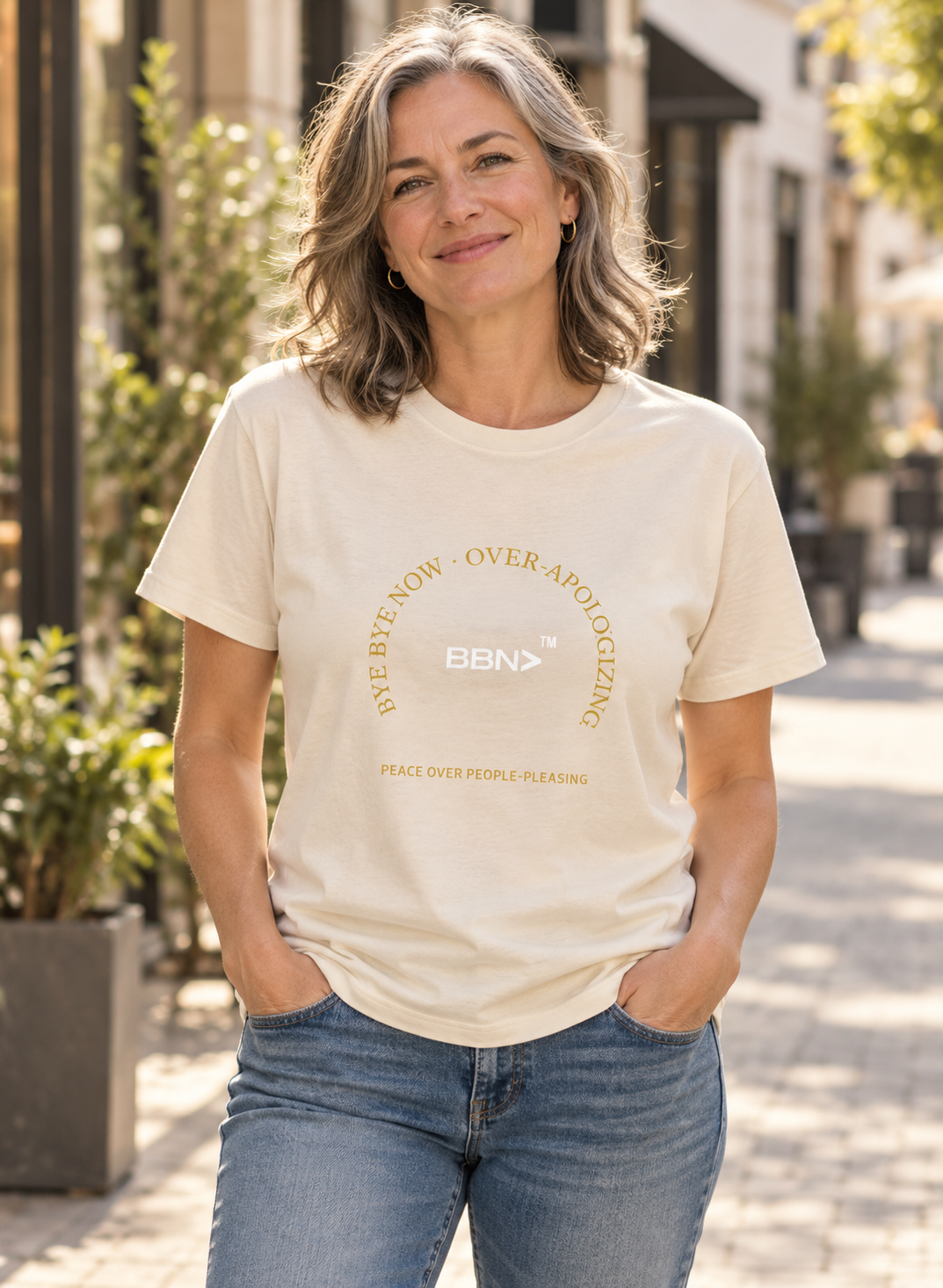 AI styling concept: Woman outdoors wearing a natural cream T-shirt with gold “Over-Apologizing” lettering around a white BBN logo.